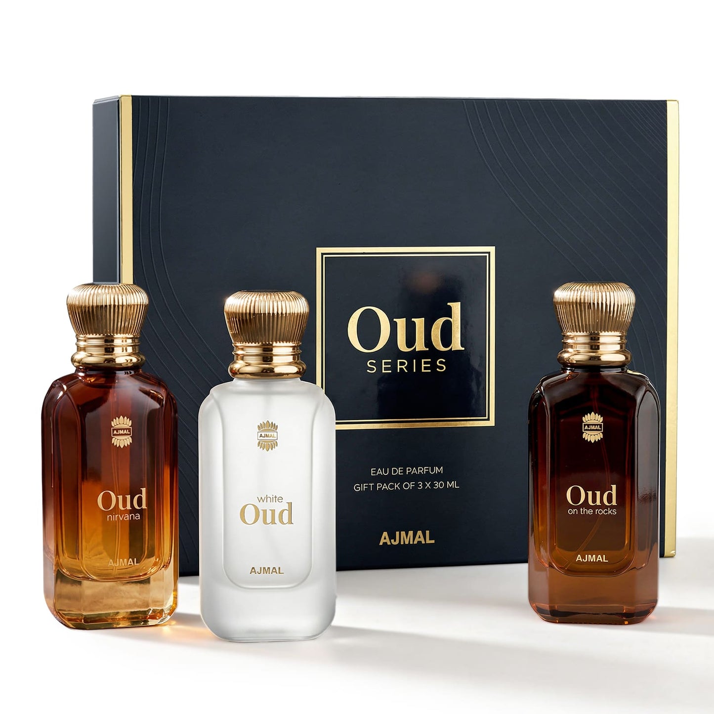 AJMAL Oud Series gift set featuring three fragrance bottles in front of a dark navy blue box with gold trim. The set includes two amber-brown bottles and one frosted white bottle, all with ribbed gold caps and gold lettering.